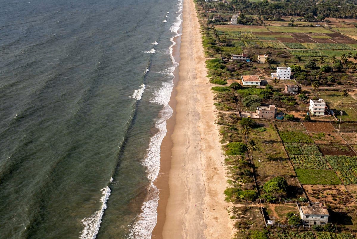 The coast from above: a long straight beach, white surf, and cultivated fields and houses along the landward side.