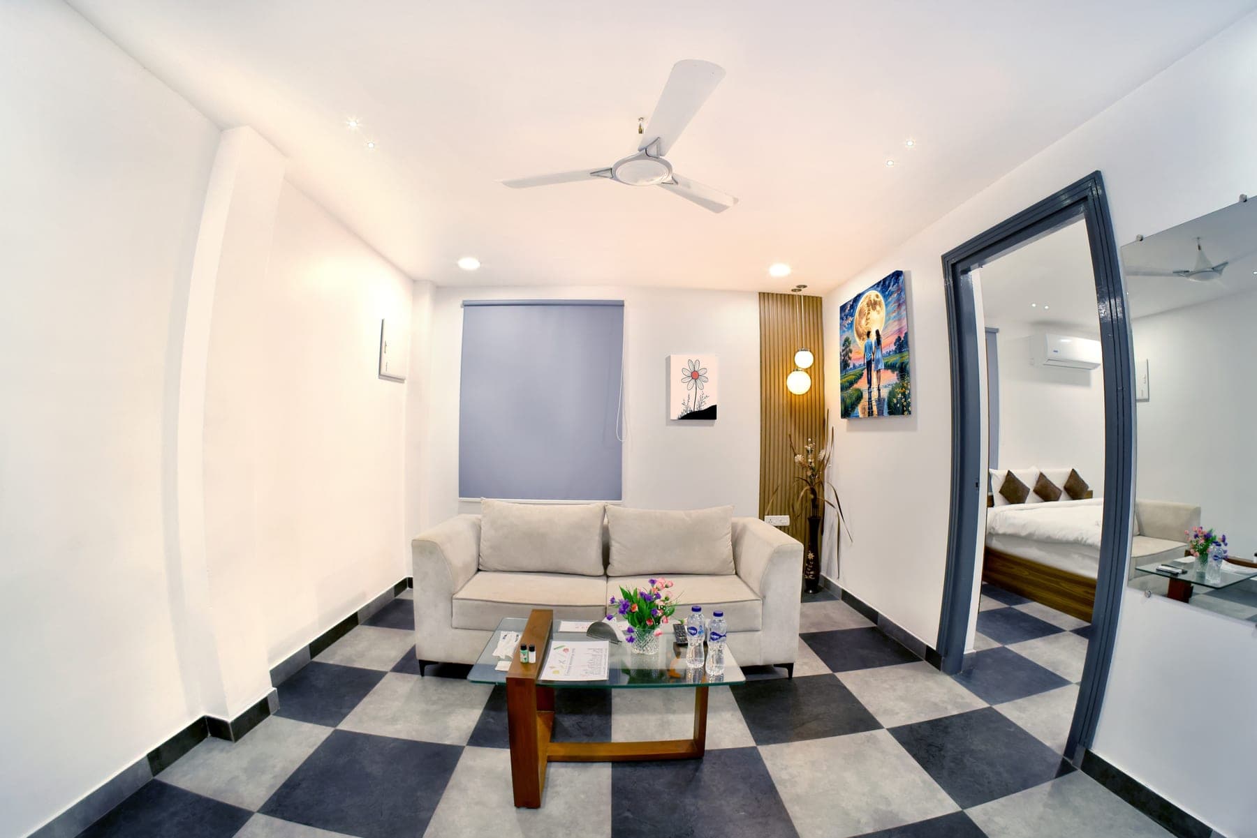 The living area of a Deluxe Cottage: a cream three-seater sofa and glass coffee table on a chequered stone floor, with an archway through to the bedroom beyond.