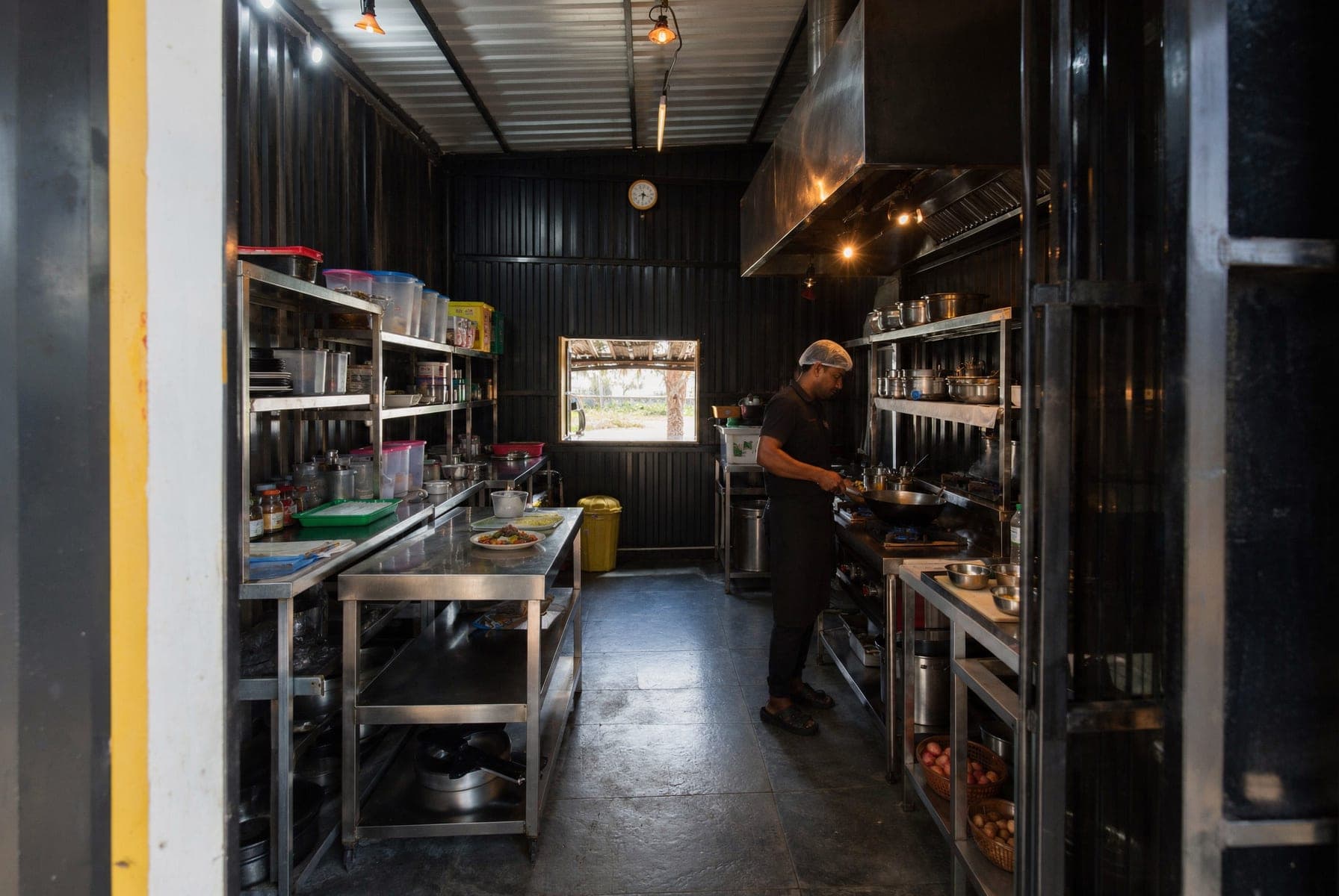 The open kitchen down its length: stainless counters and shelves of stock along both walls, a cook working a wok under the extraction hood.