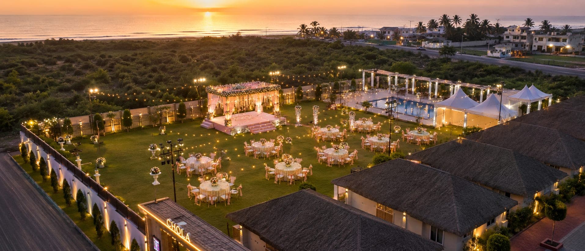 The event lawn from the air at sunset, set for a wedding: a flower-topped mandap, round tables under festoon lights, the pool and canopies to one side and the sea beyond.