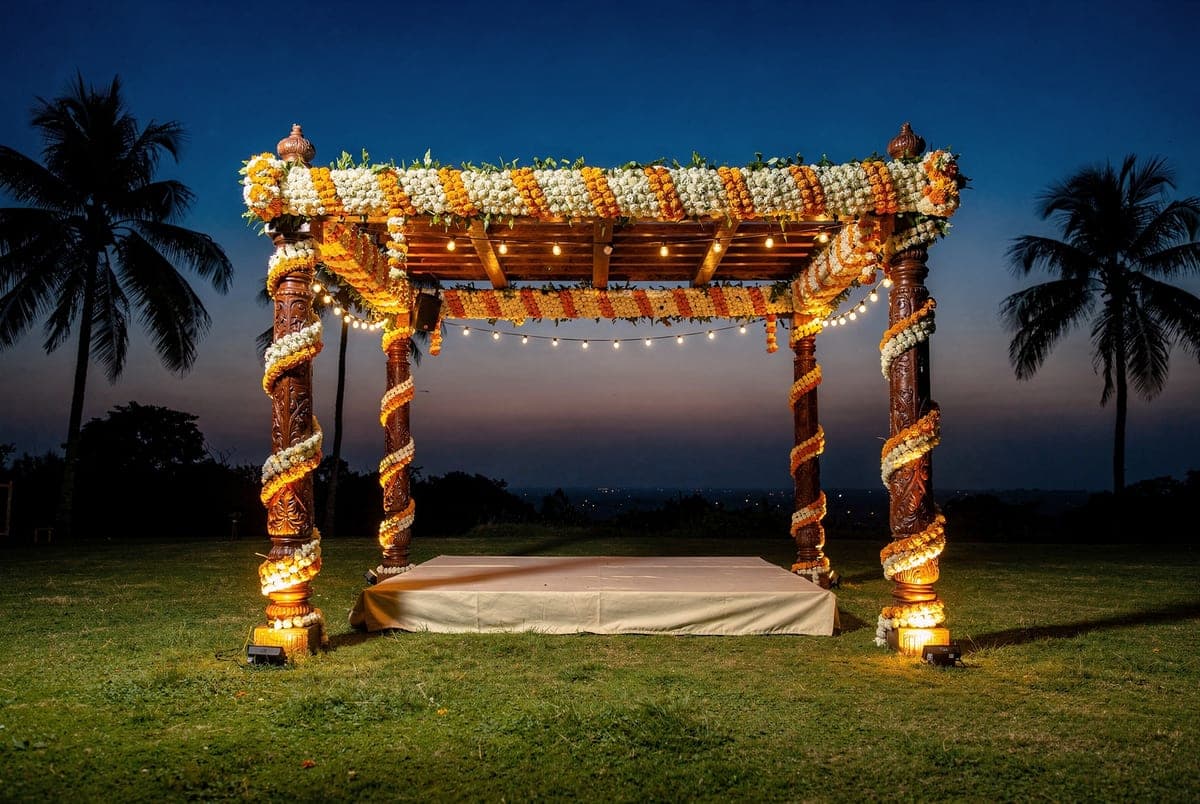 A garlanded wooden mandap lit on the lawn at dusk, marigold and white flowers wound around its four posts.