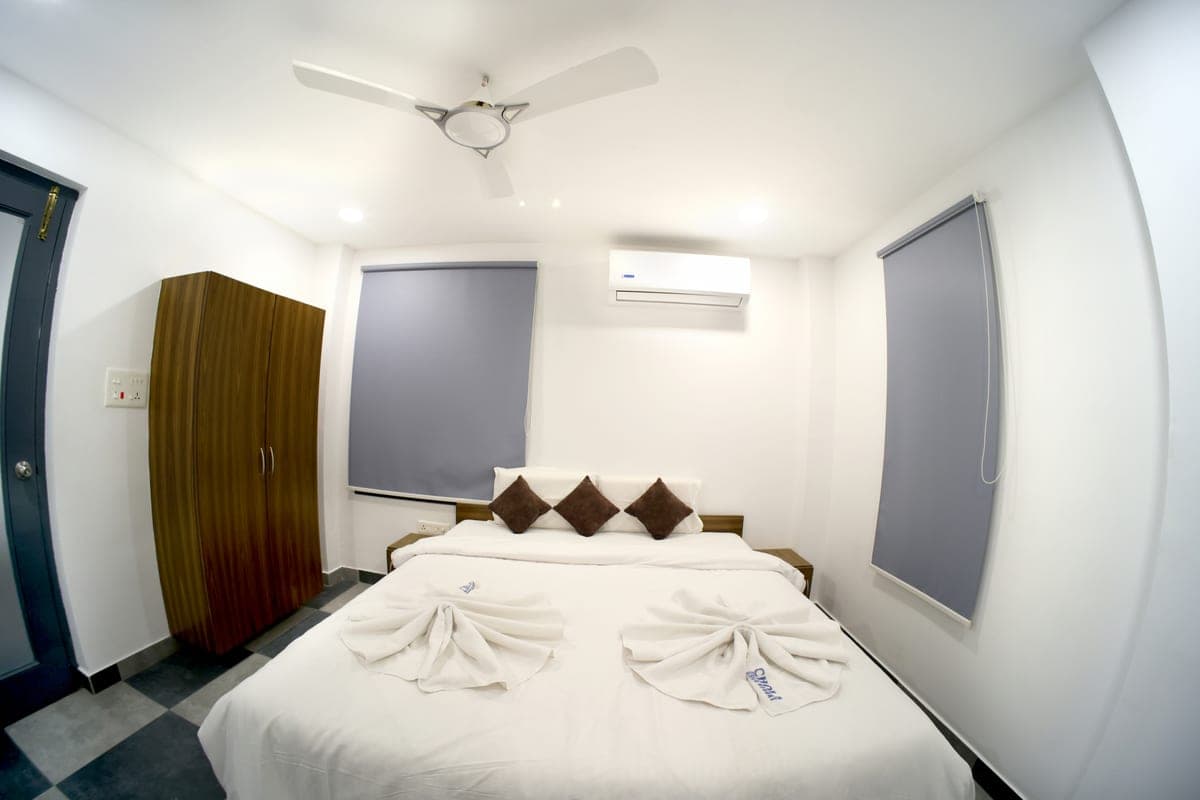 A cottage bed seen from the foot, dressed in white linen with folded towels, a wooden wardrobe alongside and an air-conditioning unit on the wall.
