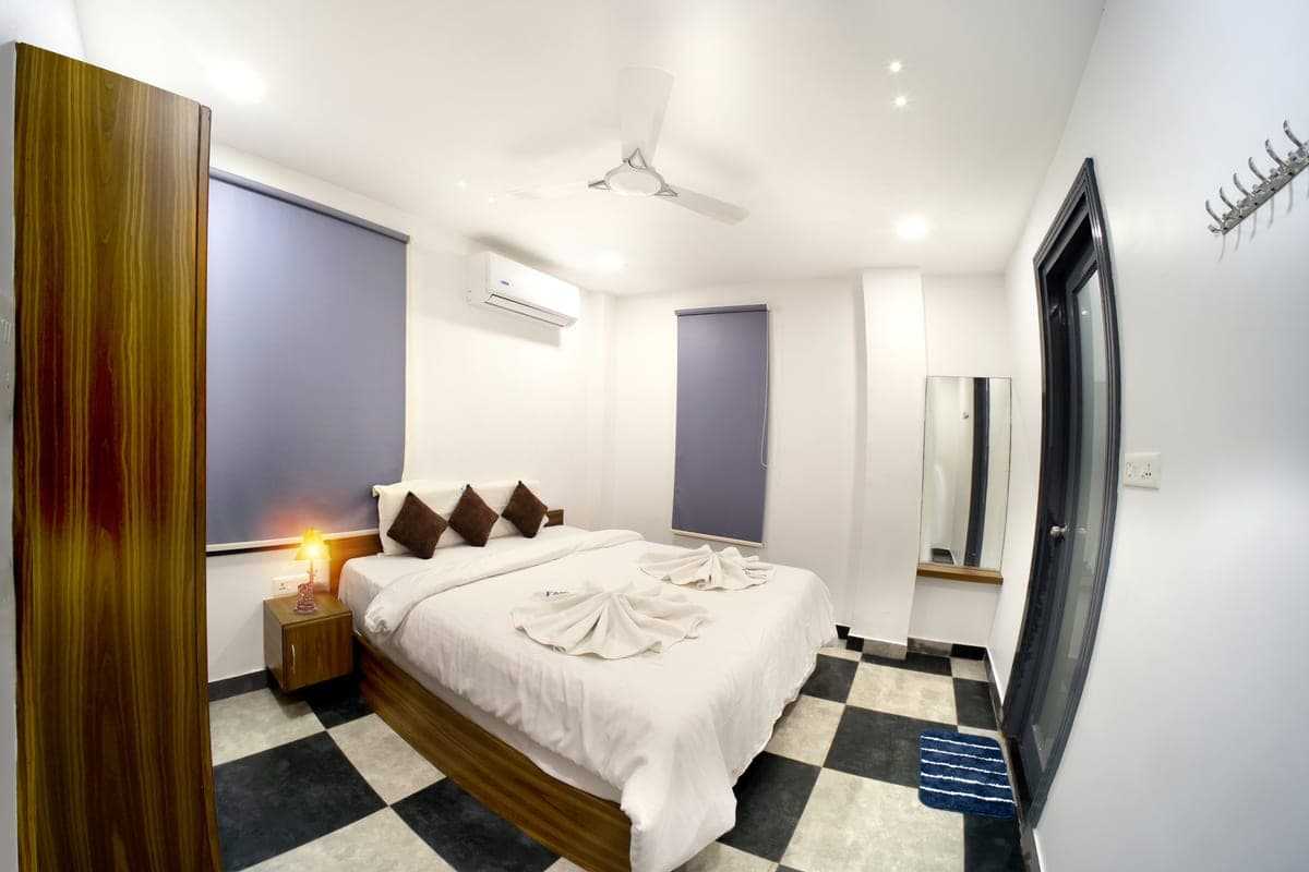 A cottage bedroom with a tall wooden wardrobe, a wide bed dressed in white linen, an air-conditioning unit above the headboard and a full-length mirror.