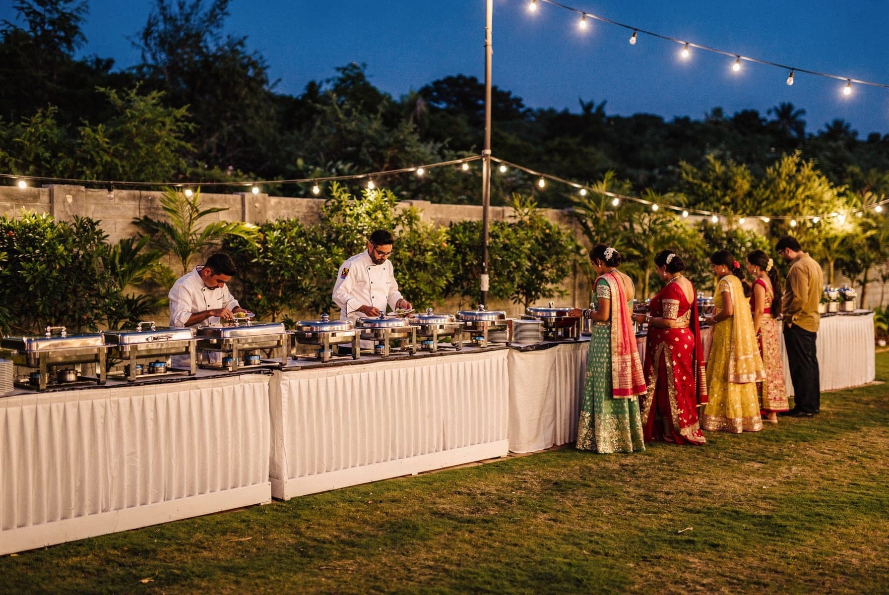 A buffet along the edge of the lawn at dusk: chefs serving from white-clothed counters of chafing dishes, guests queuing in festive clothes under festoon lights.