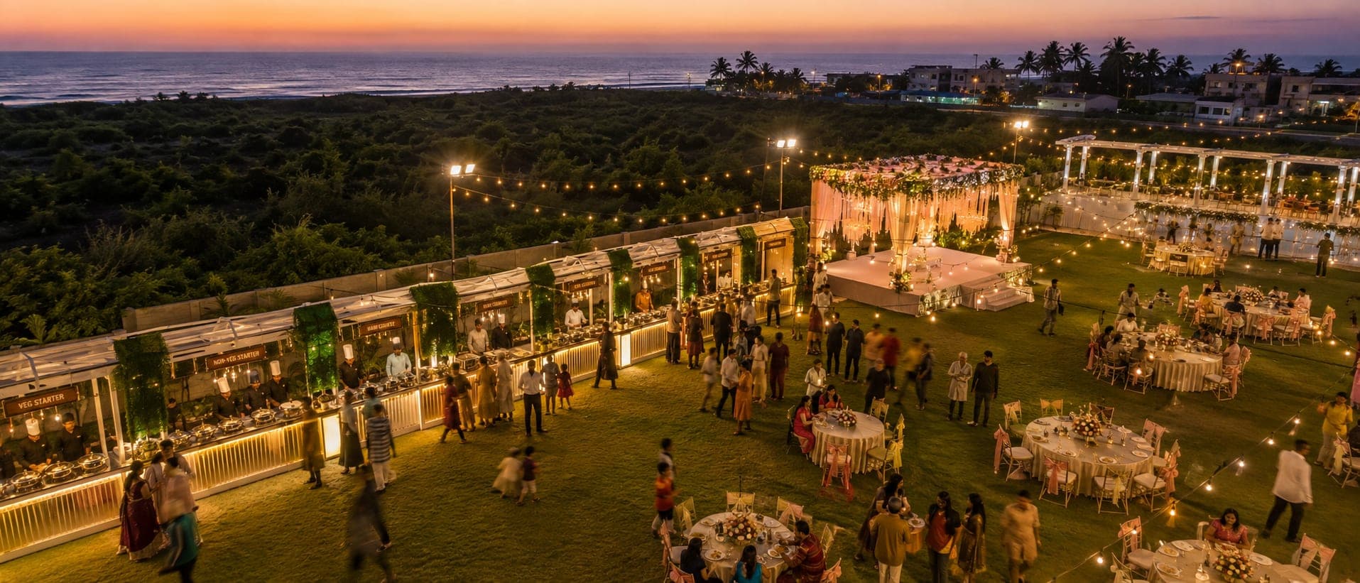 Live food counters lit along the edge of the lawn at dusk, guests moving between them, with the mandap and banquet tables beyond and the sea on the horizon.