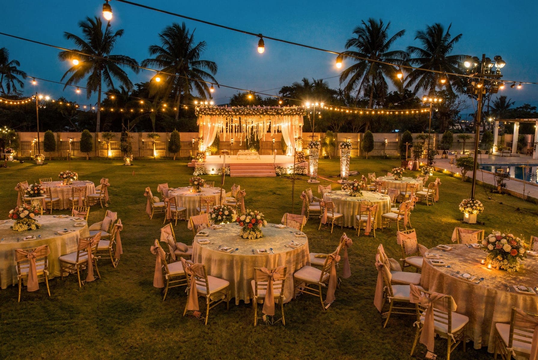 The event lawn set for dinner at dusk: round tables with gold chairs, a lit floral mandap at the far end and festoon lights overhead.