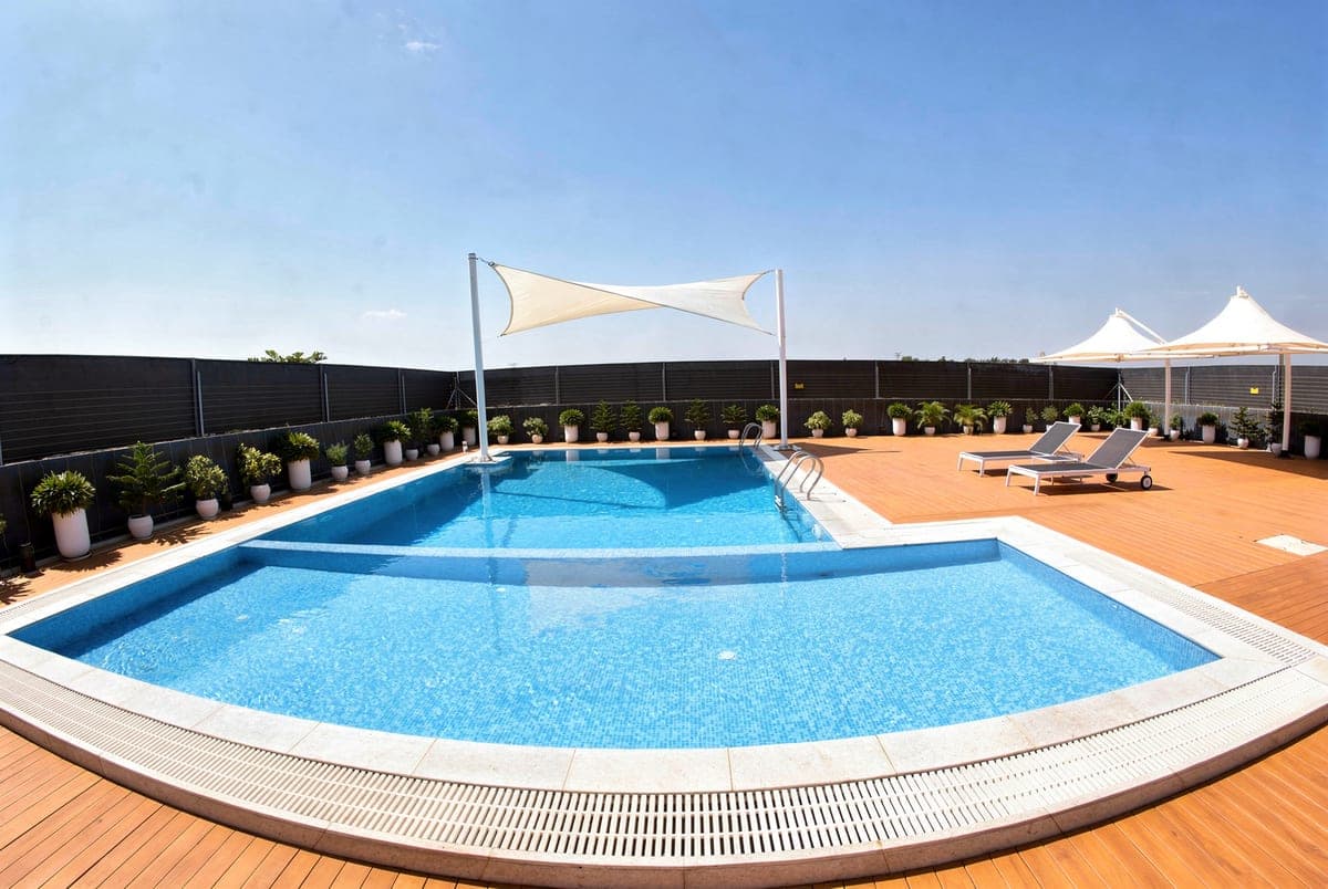 The swimming pool in bright midday sun, clear blue water, timber decking and loungers under the shade canopy.