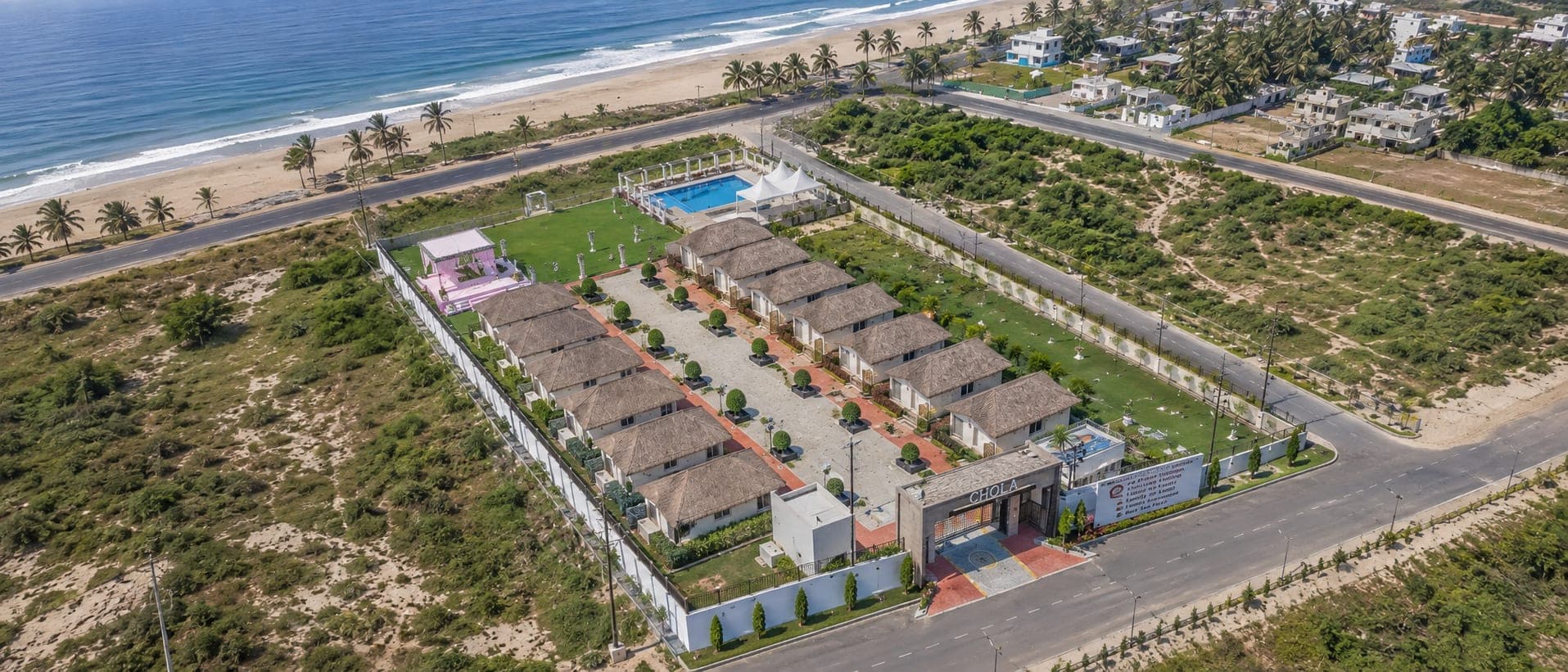 The whole resort from the air: fourteen thatched cottages in two rows, the pool and event lawn behind them, and the beach and the Bay of Bengal beyond the coast road.