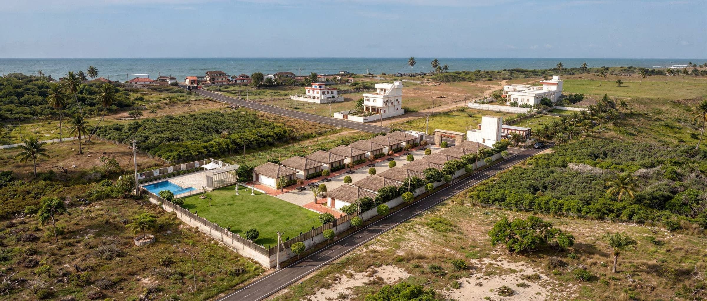 The resort from the air on a clear morning: fourteen thatched cottages in two rows inside a white-walled compound, the pool and event lawn at one end, and beyond the coast road the fields, village houses and open sand of Kothapatnam Beach.