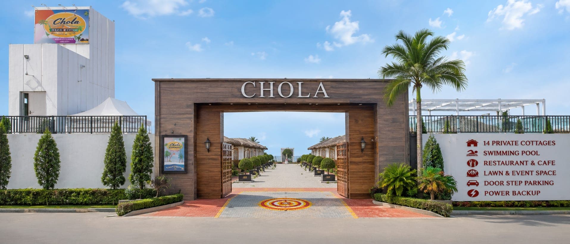 The Chola entrance gate in dark timber under a clear sky, flanked by clipped hedges and a board listing fourteen private cottages, swimming pool, restaurant, lawn and event space, doorstep parking and power backup.