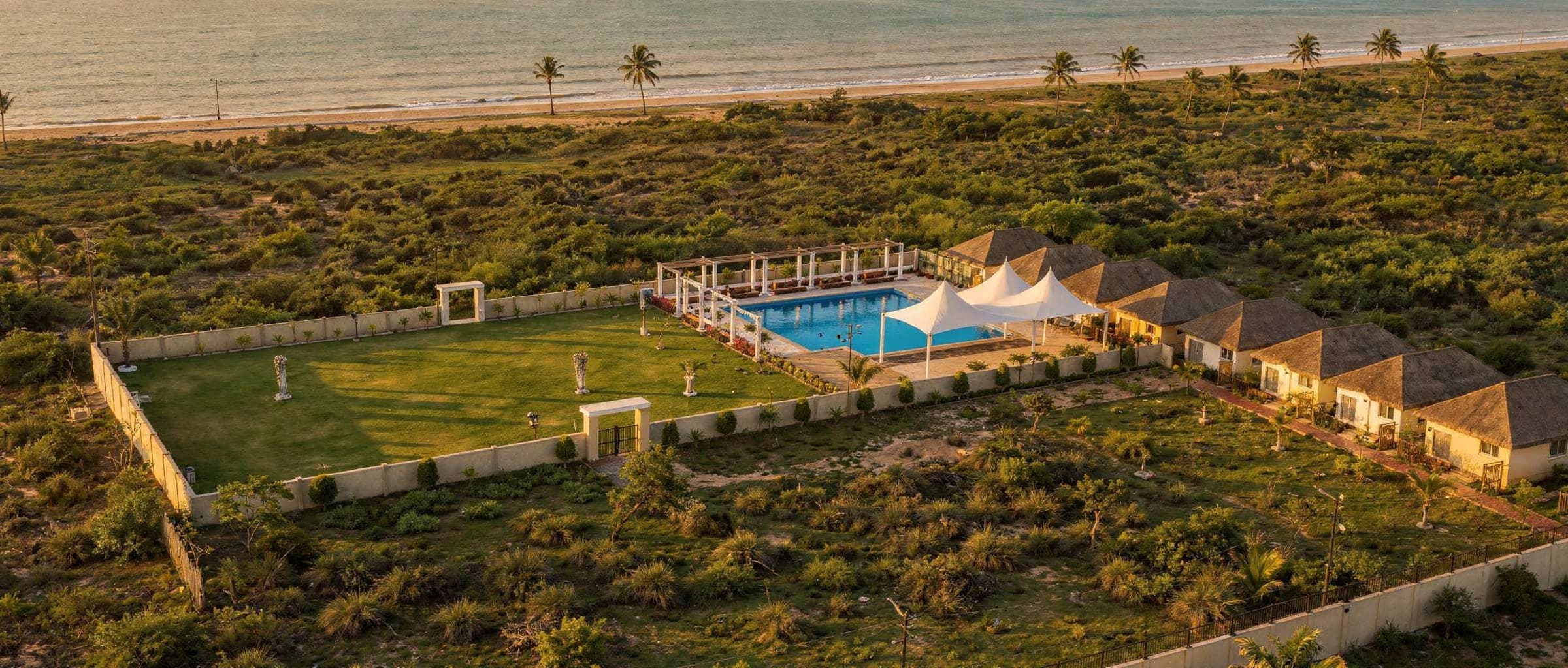 The event lawn and pool from the air at golden hour, the cottages along one side and the beach beyond the compound wall.